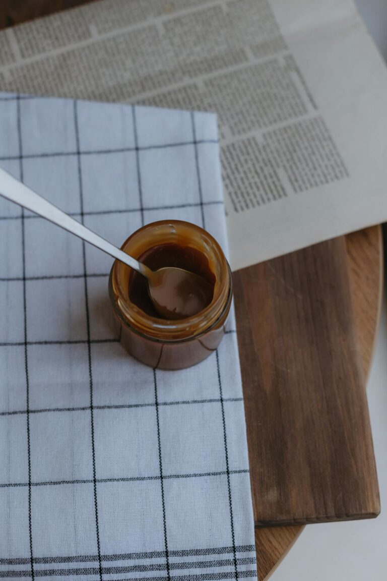 Top view of caramel sauce jar with spoon on checkered cloth atop wooden board.